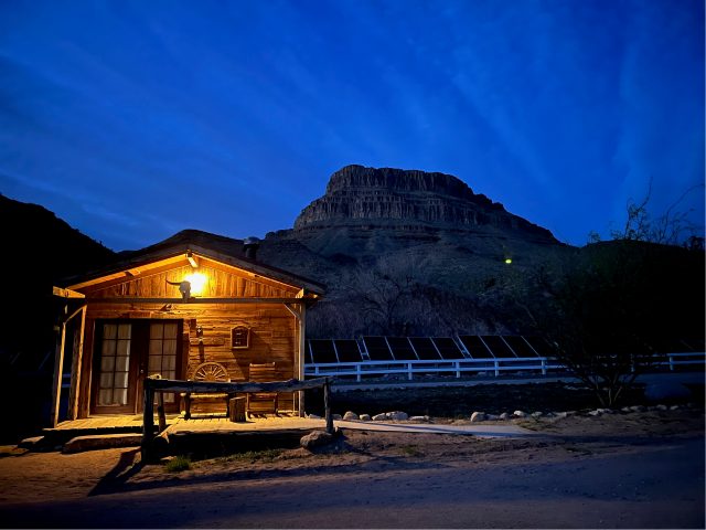 A pine cabin lit from inside at night below the dark outline of the mesa
