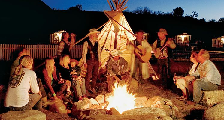 Campfire burning beside canvas tipis with guests gathered around in the dark